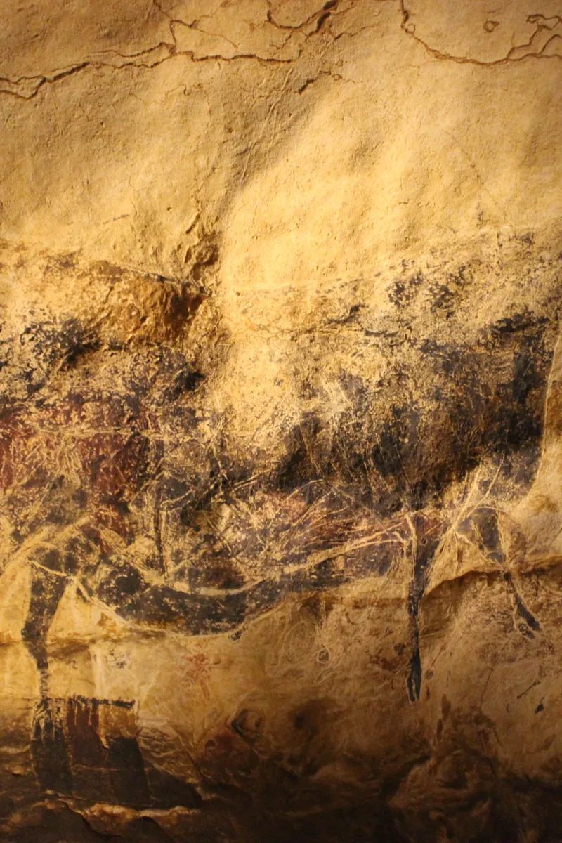 The Hall of the Bulls recreated at Lascaux IV — a curving cave wall covered in great ochre and black aurochs, horses and deer flowing across the rock, lit low in the darkness. Montignac, Dordogne, France.